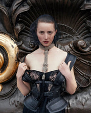 Woman in a black outfit wearing silver Elohim T-Choker necklace against a black, gold and bronze background