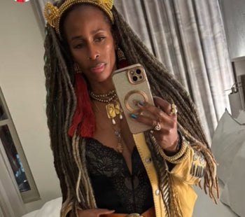 A black woman with long braided hair wearing a gold jacket and a jas headband in gold