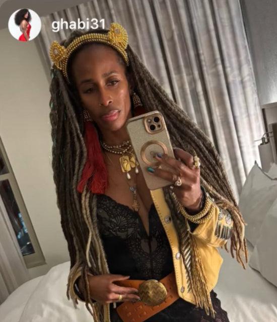 A black woman with long braided hair wearing a gold jacket and a jas headband in gold