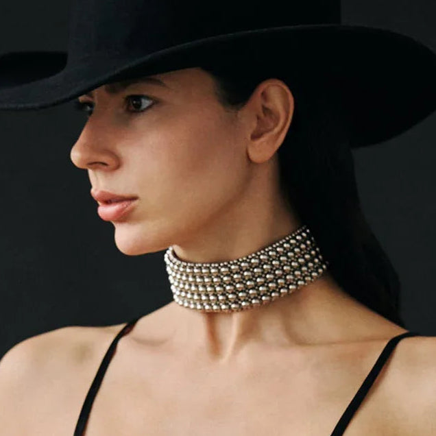 Woman wearing a black hat and black dress with a silver choker against a dark background