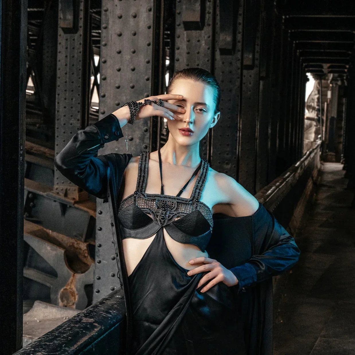 Fair skinned model wearing beaded black top and hand piece by Object & Dawn in a black silk dress standing in a dark, industrial setting