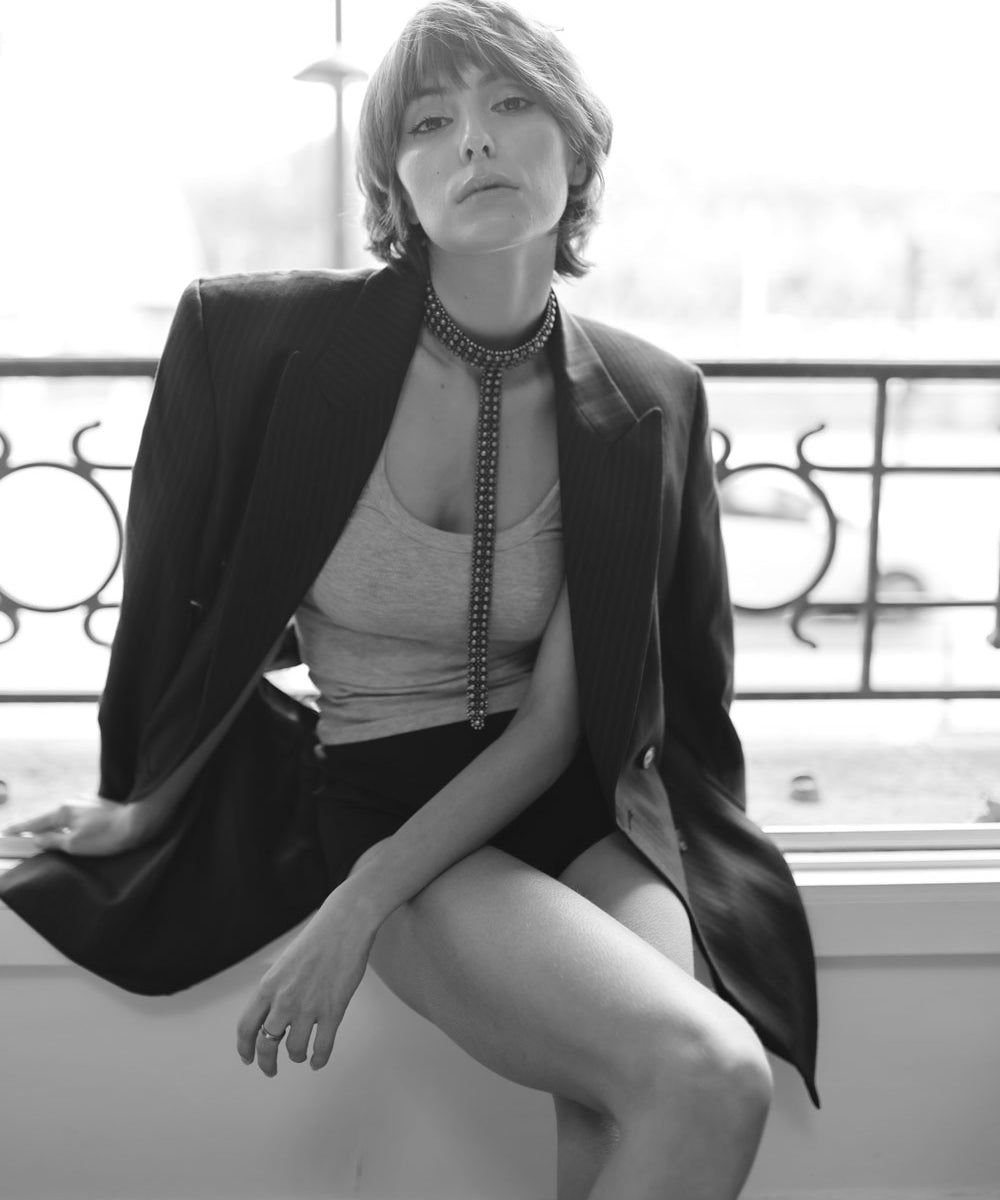 Woman wearing a black blazer and slim t-choker necklace by object & Dawn sitting on a ledge.