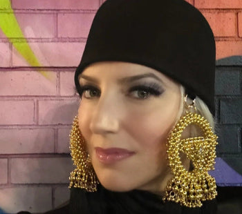 Woman wearing a black hat with gold tassels hanging on either side with a colorful brick wall background