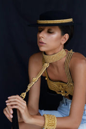 Woman wearing gold beaded jewelry by Object & Dawn and a black hat against a dark background