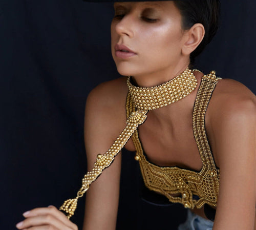 Woman wearing gold beaded jewelry by Object & Dawn and a black hat against a dark background